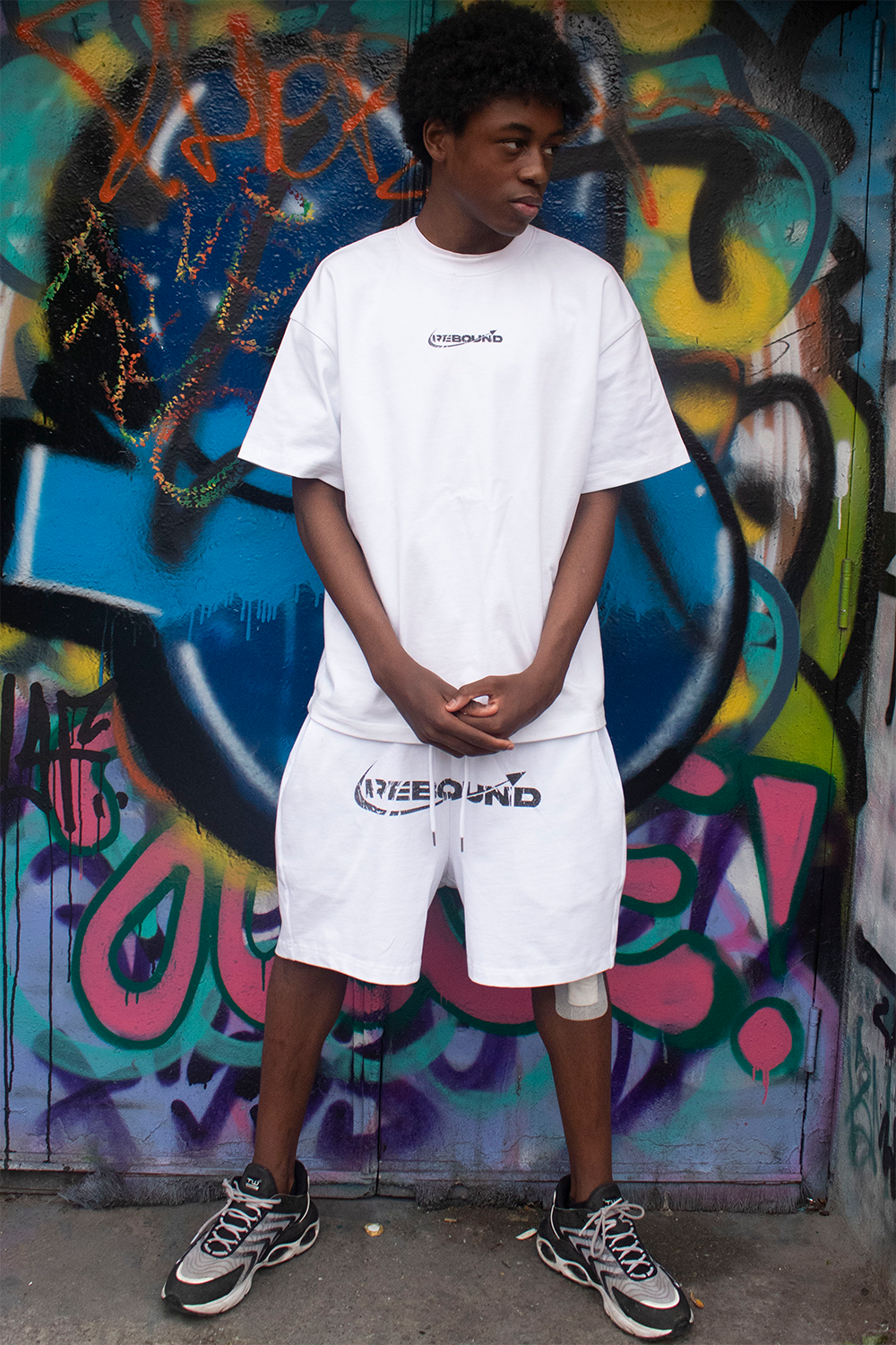 Model wearing Rebound white logo t-shirt, looking down, shot against colorful graffiti wall